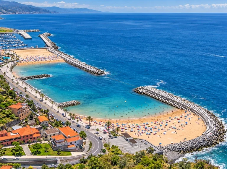 Praia da Calheta, com areia, quebra-mares e água azul junto à costa.