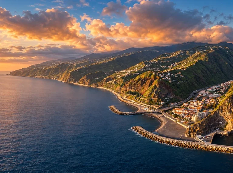 Vista da costa de Ribeira Brava ao pôr do sol, com montanhas e casas junto ao mar.